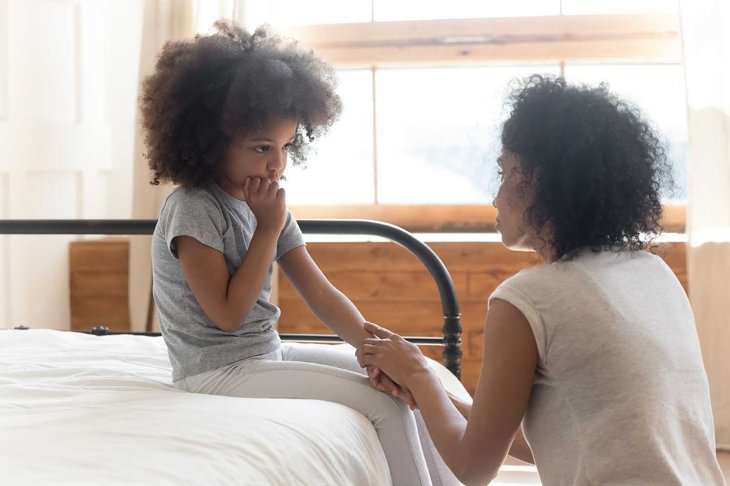 Caring worried african american mother holding hand of sad little mixed race daughter talking giving support and comfort, black mom foster parent consoling small kid being bullied sit on bed at home