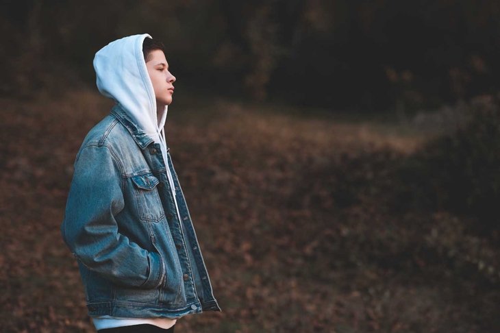Handsome teen boy wearing denim jacket and white hoodie standing outdoors over nature autumn background. Teenagerhood.